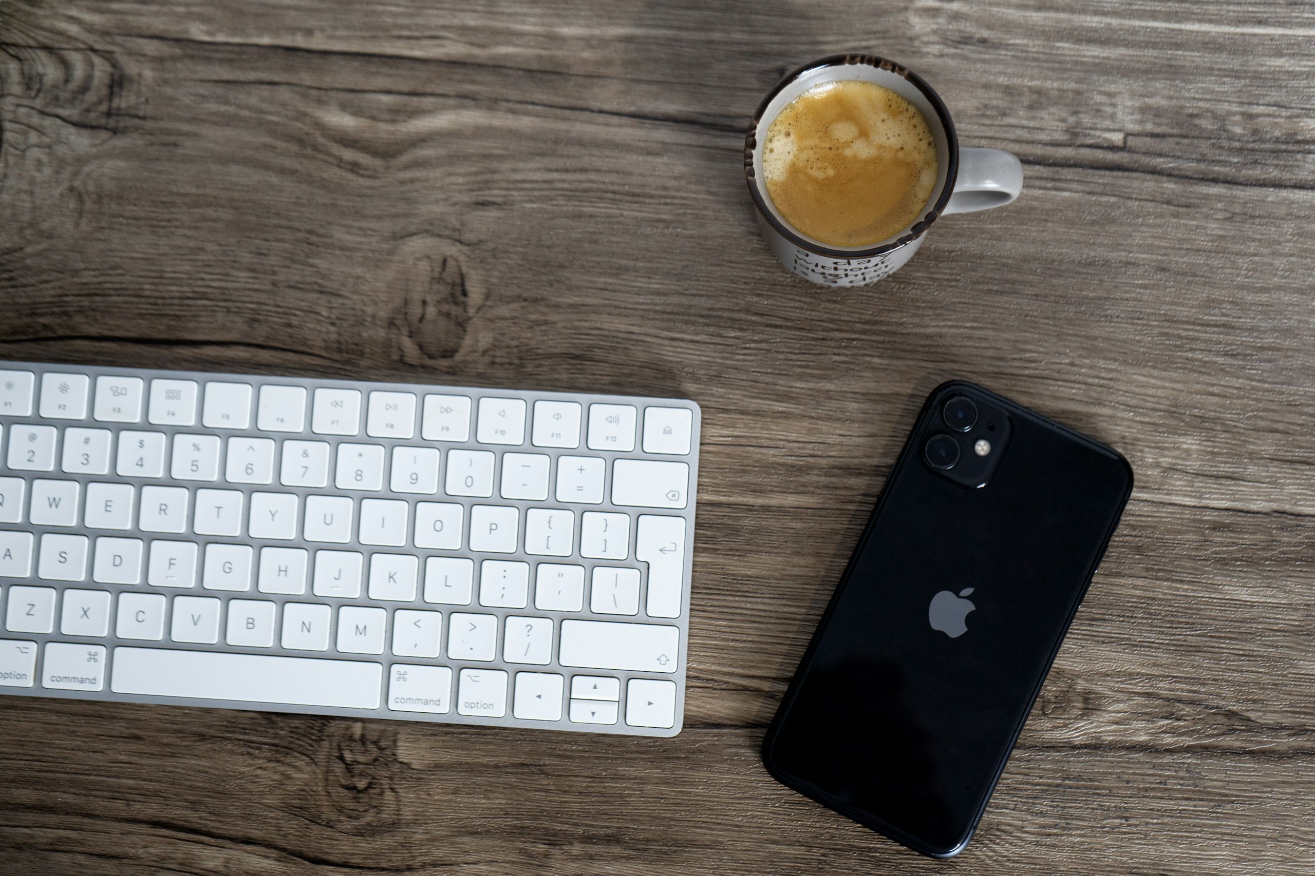 a black smartphone, white keyboard, and a cup of coffee arranged on a wooden desk